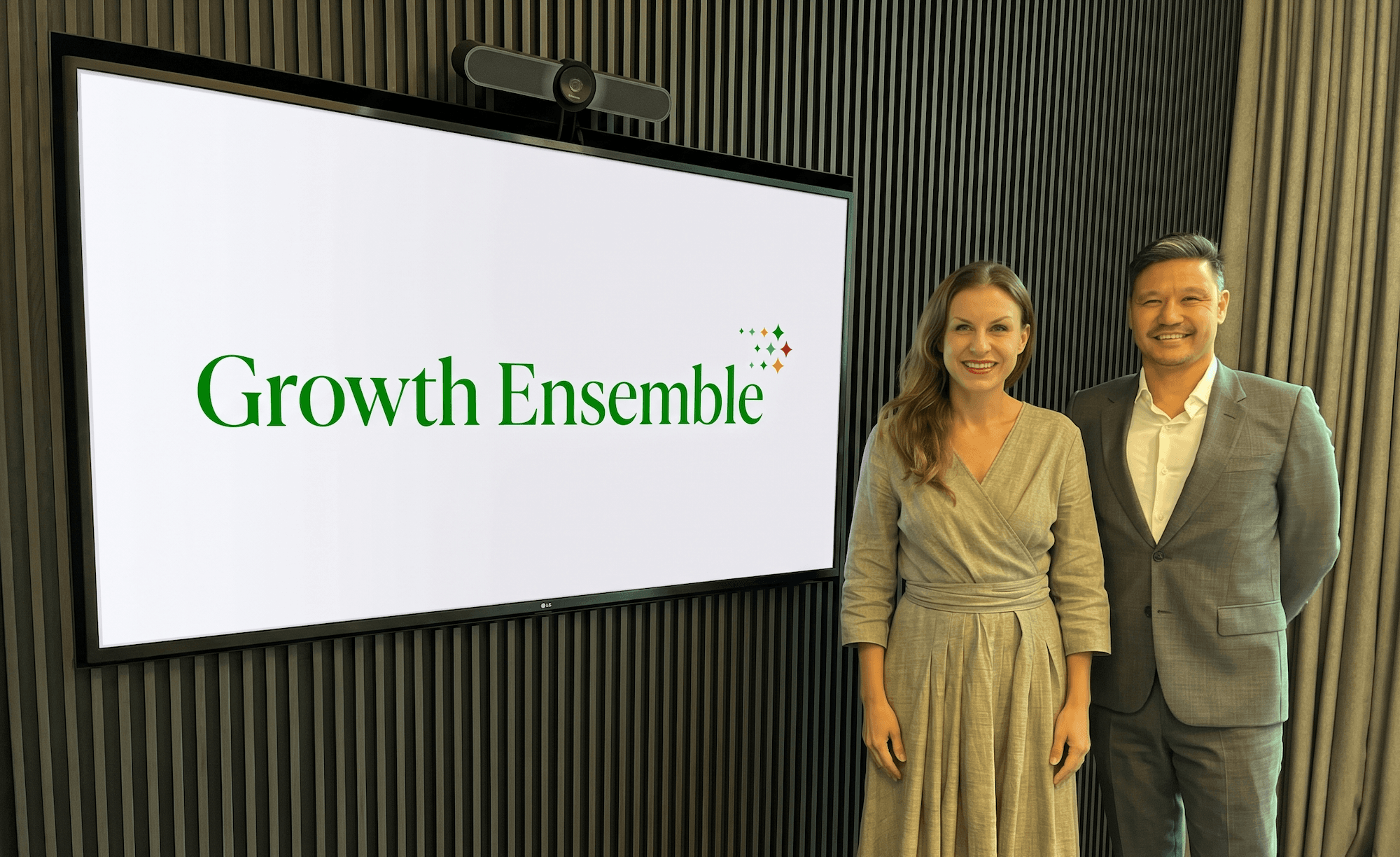 Growth Ensemble Co-founders and Managing Partners Meredith Carson and Adam Flinter standing next to a TV screen with their company logo on it.
